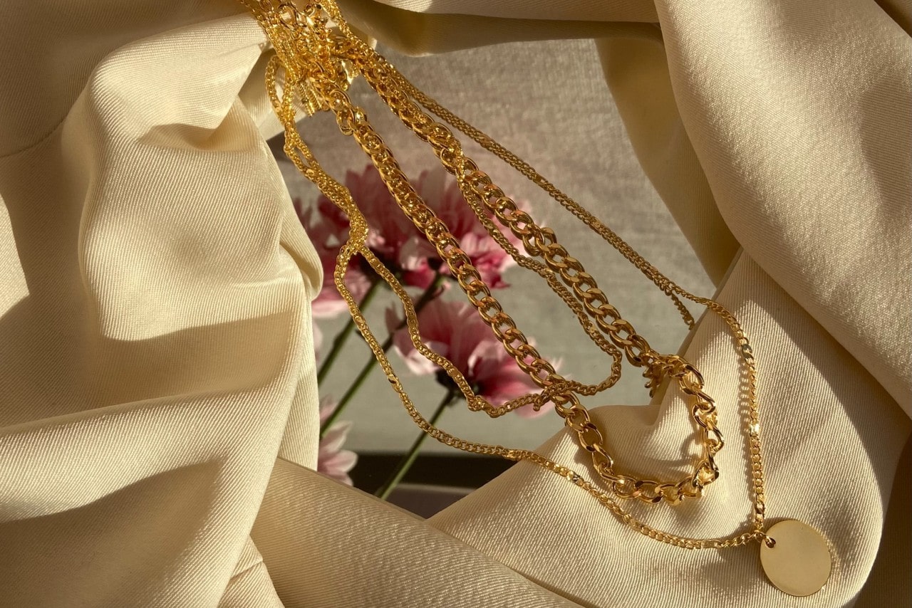 A trio of chain necklaces sit on a mirror and cream linen.