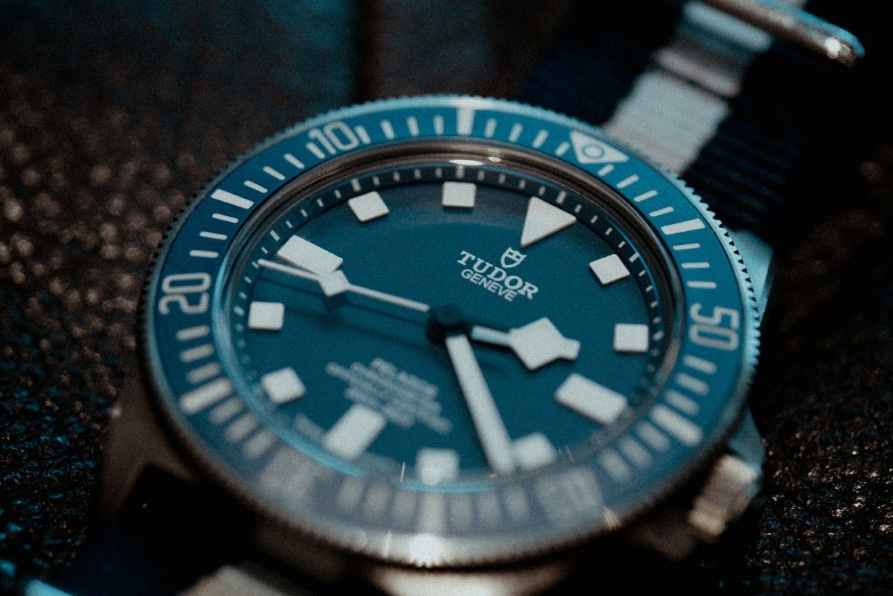 A close-up of a TUDOR Pelagos watch with a blue dial and fabric strap on a textured backdrop.