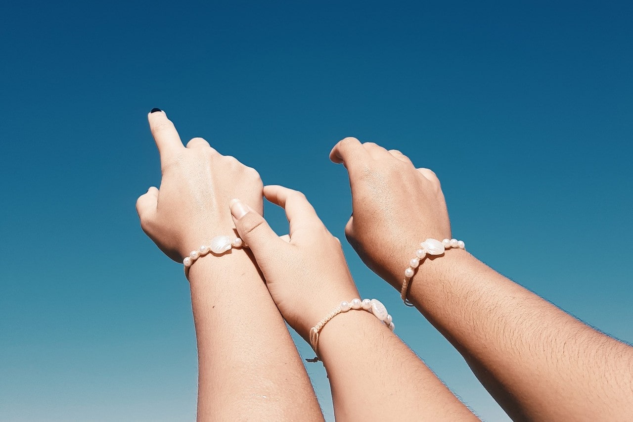 Three women reach toward the sky, wearing matching seashell bracelets.